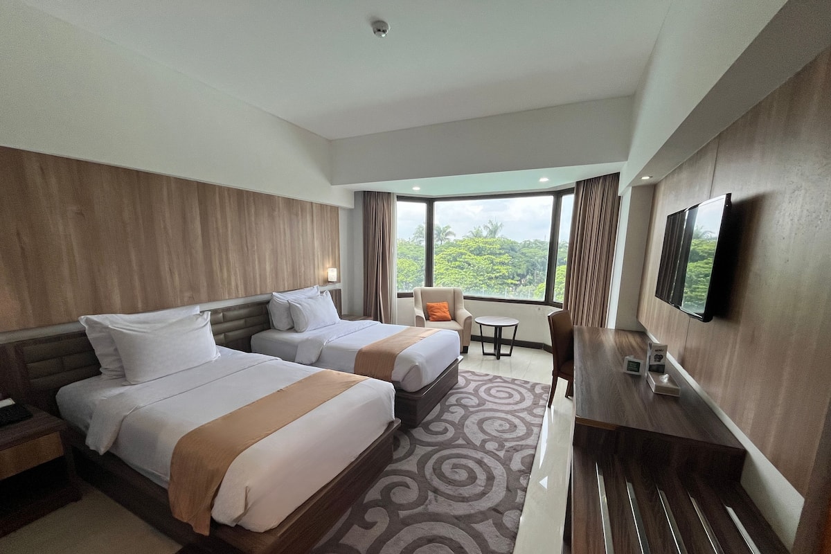 A hotel room with two beds, a desk, a chair, a TV, and a large window with a view of greenery.
