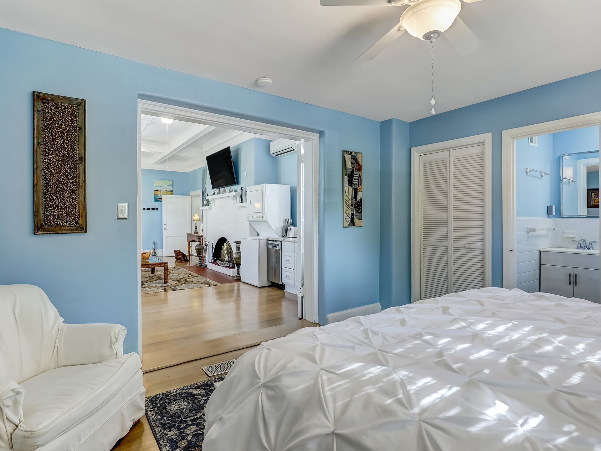 A bedroom with a bed, a white armchair, a ceiling fan, and a bathroom with a mirror and sink.