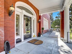 A brick building with a white door, numbered 102, and a covered porch with outdoor seating. - Walk Everywhere in Starland, Heated Pool Access (Savannah)