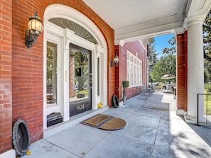 A brick building with a white door, numbered 102, and a covered porch with outdoor seating.