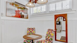 Un coin repas avec une table ronde et deux chaises, un grand tableau, un miroir et une fenêtre avec des stores.