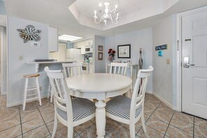 A dining area with a white round table and chairs, a chandelier, and a kitchen in the background. - Oceanfront Condominium with Amenities (Saint Simons Island)