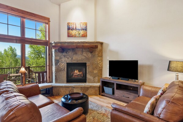 Una sala de estar con chimenea de piedra, sofás de cuero, televisor y balcón con mobiliario exterior.