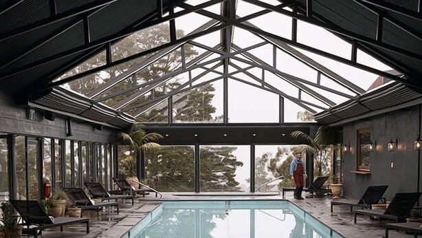 Piscina interior moderna com teto de vidro, espreguiçadeiras e plantas em vasos.