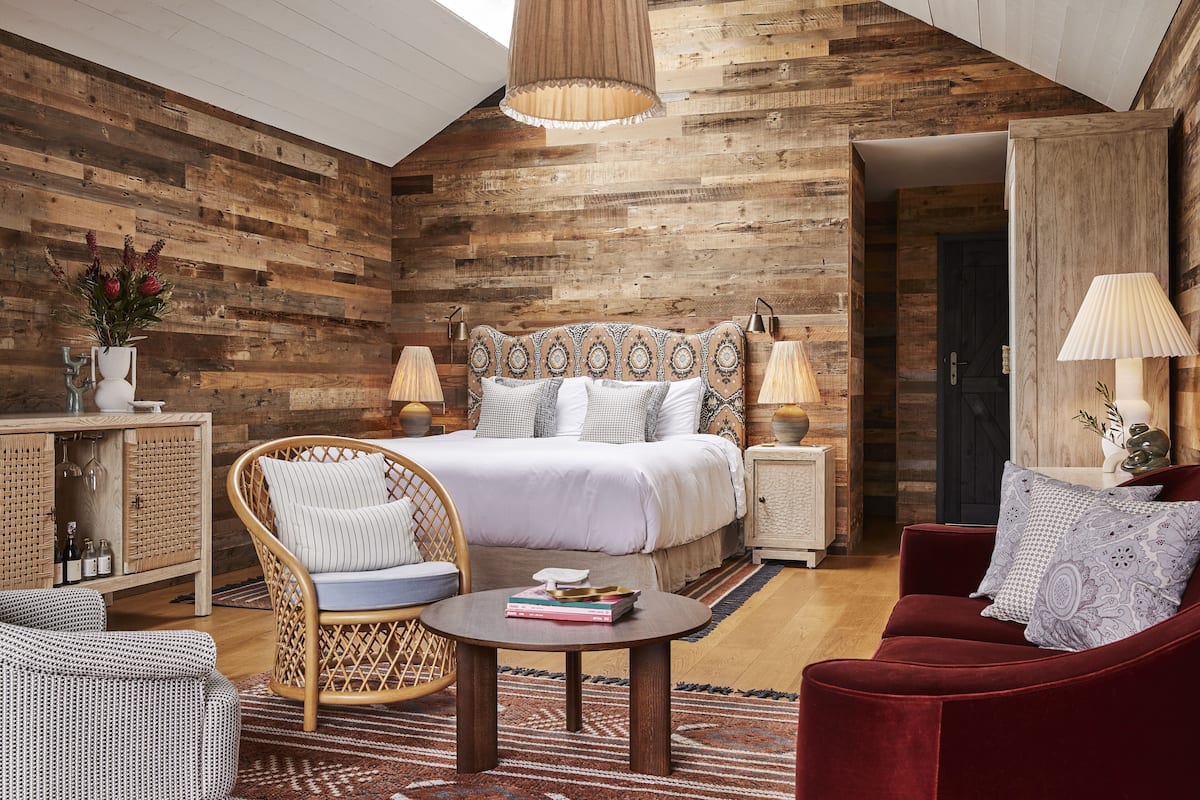 A cozy bedroom with a wooden slat ceiling, a large bed with patterned headboard, a wicker chair, a red sofa, and a small table with books.