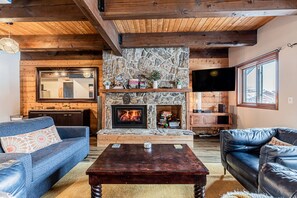 Un salon chaleureux avec une cheminée en pierre, des poutres en bois, un téléviseur et des sièges confortables.