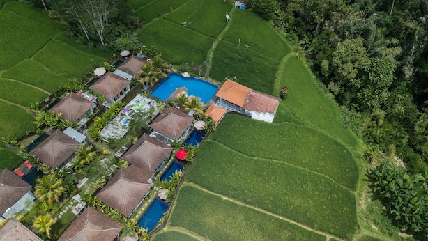 An aerial view of a resort with multiple bungalow-style buildings, a central swimming pool, and lush green fields. - Tanta Escape (Sebatu)