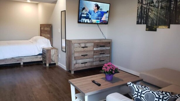 A bedroom with a bed, a wooden dresser, a TV mounted on the wall, and a small table with a remote control and a potted plant.