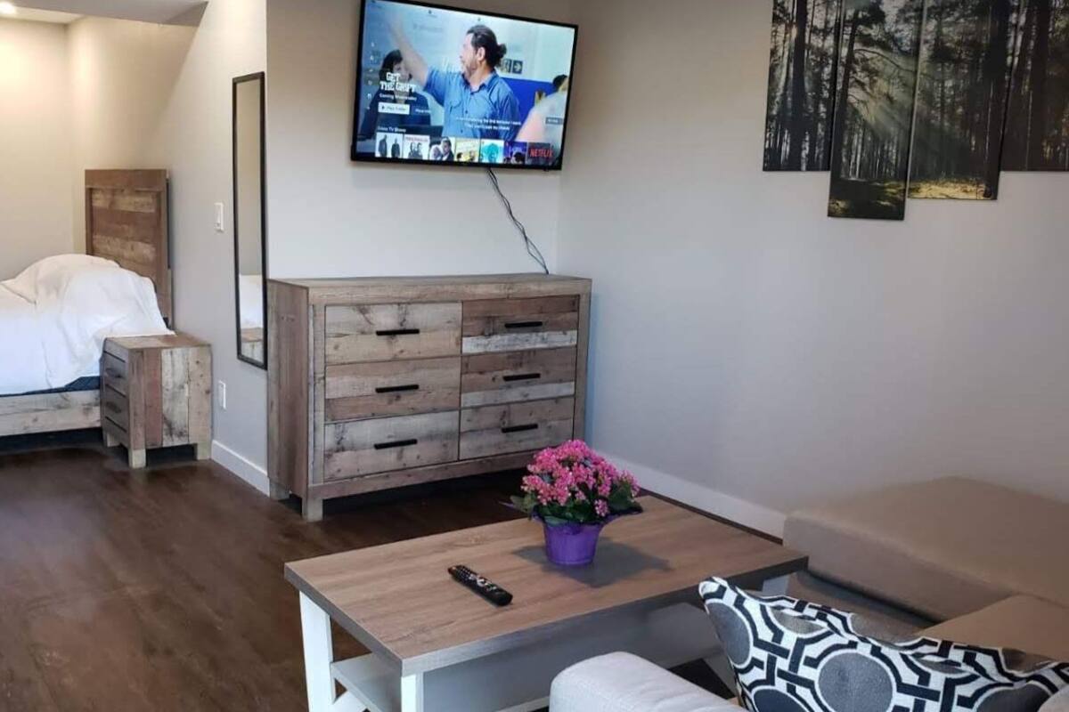 Un dormitorio con cama, cómoda de madera, televisor montado en la pared y una mesita con control remoto y planta en maceta.