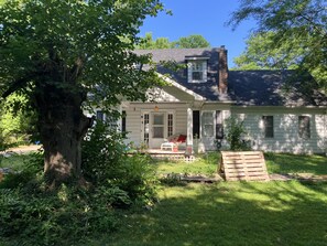 Ein Haus mit Veranda, ein großer Baum davor und eine kleine Gartenanlage.