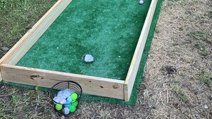 Une structure en bois recouverte de gazon synthétique, un panier en métal avec des ballons, et une clôture en bois en arrière-plan.