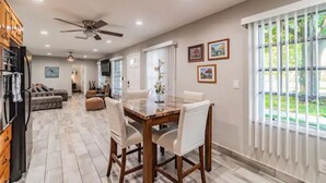 A spacious dining area with a glass-top table and white chairs, a ceiling fan, and a view of a garden through large windows. - Town 'n' Country Cozy Corner: Your Florida Fun Hub (Tampa)