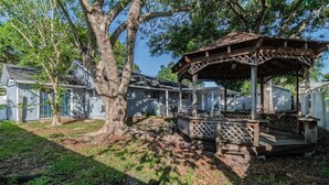 A gazebo with a wooden lattice design, surrounded by a grassy area with trees and a house in the background. - Town 'n' Country Cozy Corner: Your Florida Fun Hub (Tampa)
