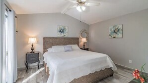 A bedroom with a large bed, two bedside tables with lamps, a ceiling fan, and floral artwork on the walls. - Town 'n' Country Cozy Corner: Your Florida Fun Hub (Tampa)
