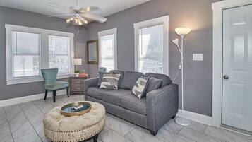 Un salon avec un canapé gris, un fauteuil turquoise et un pouf rond. Il y a trois fenêtres équipées de stores, un ventilateur de plafond et une lampe sur pied.