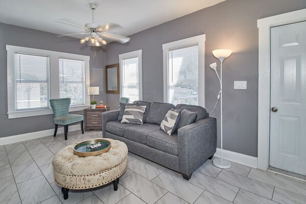 A living room with a gray sofa, teal armchair, and a round ottoman. There are three windows with blinds, a ceiling fan, and a floor lamp.
