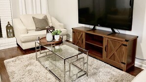 Een woonkamer met een witte bank, een glazen salontafel, een houten tv-meubel en een grote flatscreen televisie.