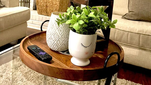 Een woonkamer met een wit bakstenen haardvuur, een ronde houten salontafel met een potplant, een afstandsbediening, en een geweven mand.