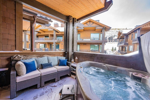 A wooden balcony with a hot tub, a sofa with cushions, and a view of other buildings.