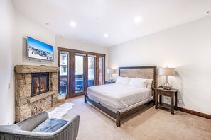 A bedroom with a stone fireplace, a large bed, a nightstand, and a view through a glass door. - Juniper Landing (Park City)