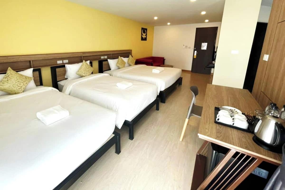 Una habitación de hotel con tres camas, un cabecero de madera, una mesa de comedor con un hervidor y una silla roja.