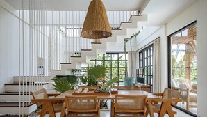 A spacious dining area with a high ceiling, a staircase with white railings, and large windows offering views of greenery. - Rumah Simba (Munggu)