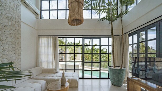 A spacious living room with a large window, a stone wall, a white sofa, a wooden bench, and a potted plant. - Rumah Simba (Munggu)