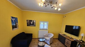 Ein Wohnzimmer mit gelben Wänden, einem schwarzen Sofa, einem weißen Sessel, einem kleinen runden Tisch, einem Fernseher und einer Kommode mit Spiegel.