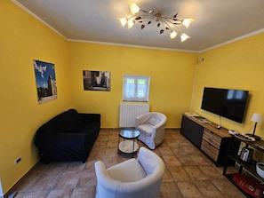 Een woonkamer met gele wanden, een zwarte bank, een witte fauteuil, een kleine ronde tafel, een televisie en een kast met spiegel.