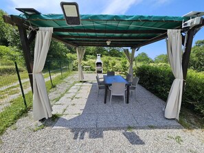 A covered outdoor seating area with a table and chairs, a grill, and a pergola structure. - The Look of Nature (Framura)