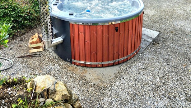 Ein Whirlpool mit Holzumzäunung, umgeben von Kies und Grünpflanzen, mit Bergen im Hintergrund.