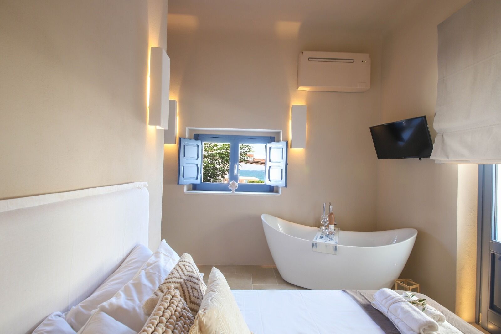 Un baño moderno con bañera, ventana con persiana abierta y televisor montado.