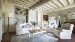 A cozy living room with a white sofa, a wooden coffee table, and a white armchair. There is a bookshelf with decorative items and a fireplace. - Villa Marraccini 8 in Lucca (Lucca)