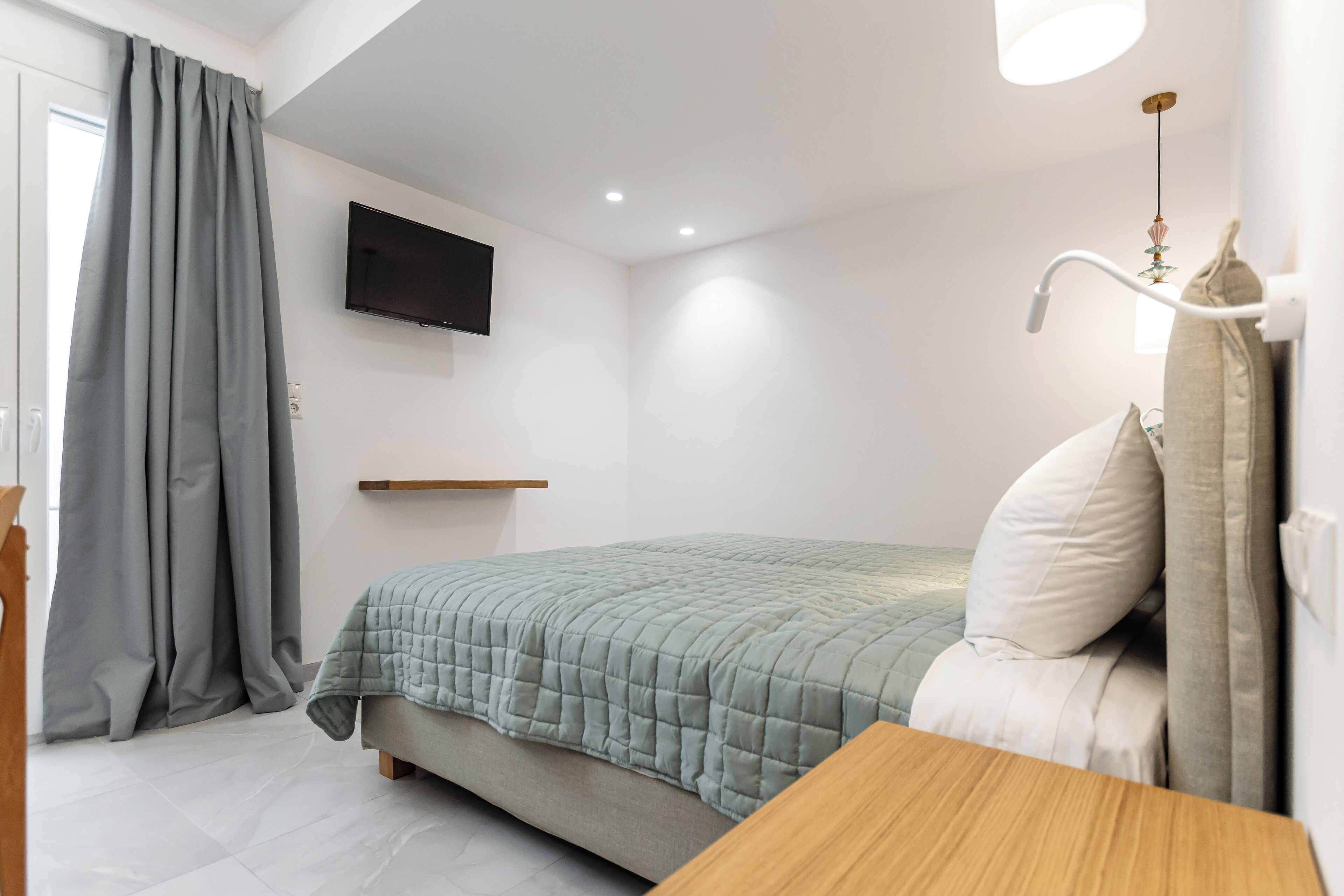 Une chambre à coucher avec un lit, une table de chevet en bois, un téléviseur fixé au mur et une fenêtre avec des rideaux.