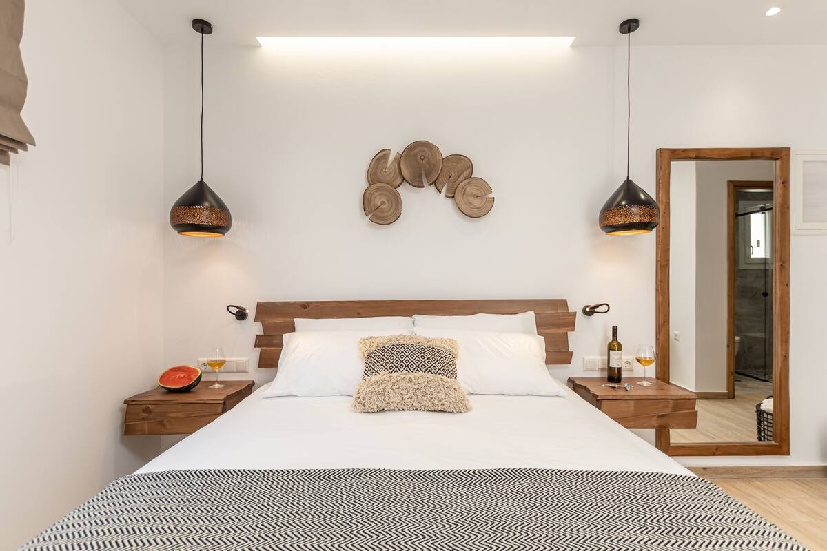 A modern bedroom with a large bed, wooden headboard, two bedside tables with wine glasses, a fruit bowl, and a mirror.