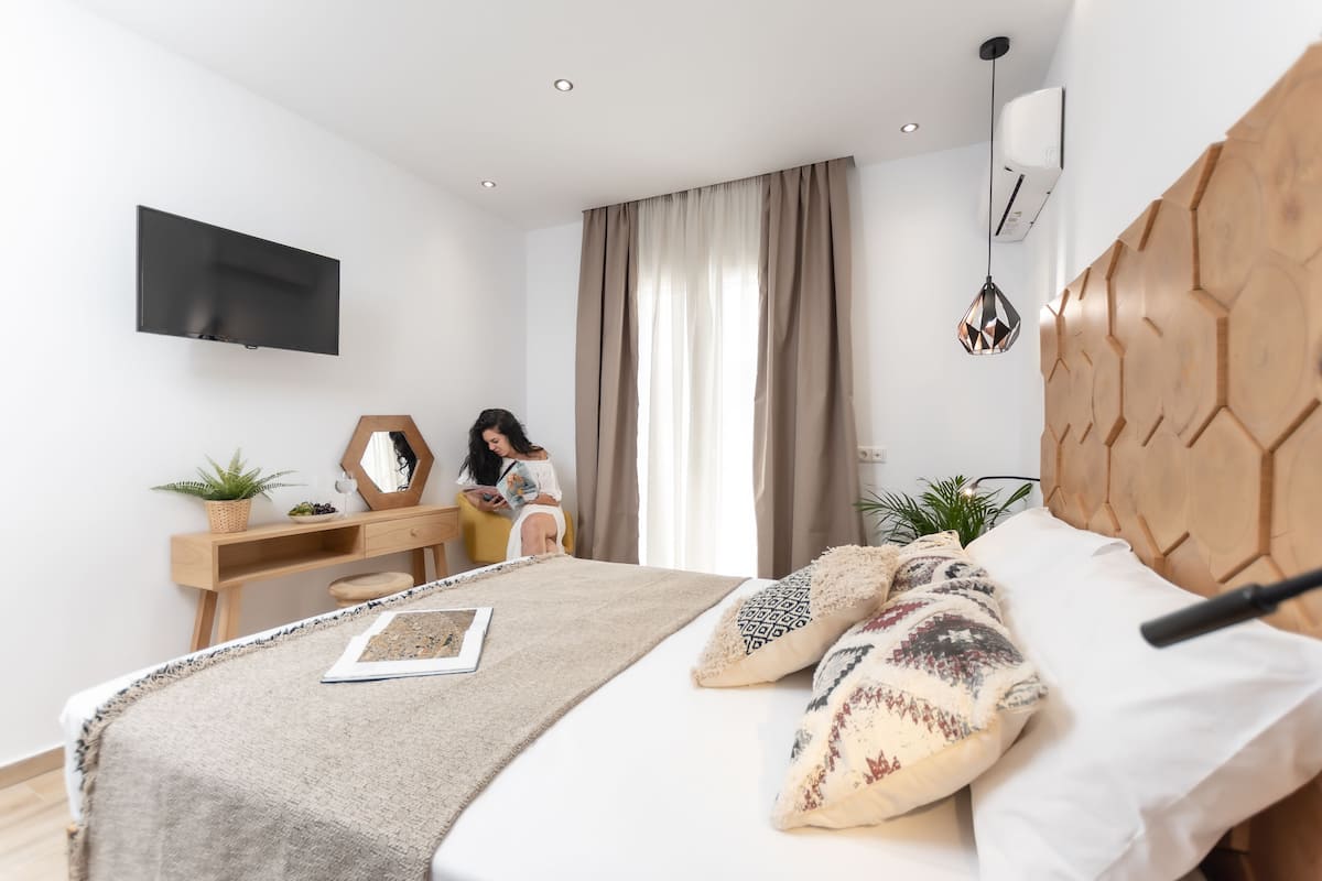 A modern bedroom with a large bed, a wooden headboard, a desk with a chair, a flat-screen TV, and a large window with curtains.