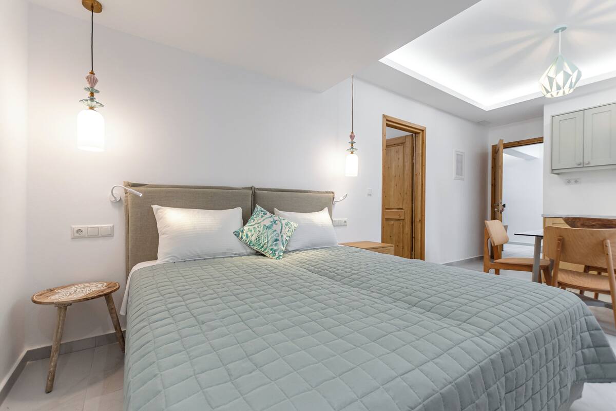A bedroom with a large bed, a wooden nightstand, a small wooden stool, a desk with a chair, and a wooden door.