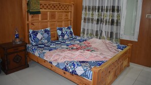 A wooden bed with a quilted headboard, a bedside table with a bottle and glasses, and a window with curtains.