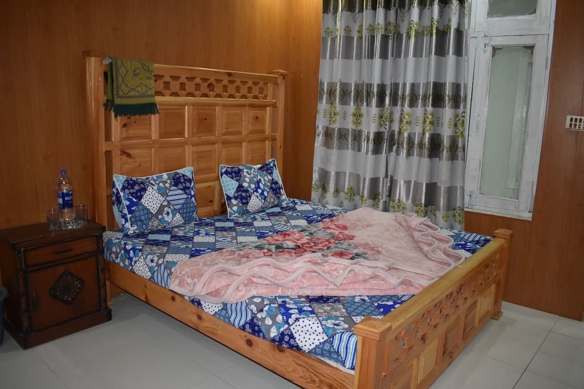 Un lit en bois avec une tête de lit matelassée, une table de chevet avec une bouteille et des verres, et une fenêtre avec des rideaux.