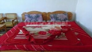 A bed with a red blanket and floral pillows, wooden headboard, and two chairs in the background. - Frontier Hotel (Balakot)
