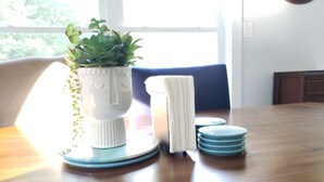 Ein Esstisch mit einer weißen Keramikvase, einer Pflanze, einem Stapel weißer Servietten und blauen Untersetzern.