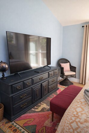 Een slaapkamer met een donker houten dressoir, een flatscreen televisie, een rieten fauteuil, een kamerkleed met dessin en een bed met een rode deken.