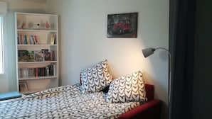Un dormitori amb un estovat estampat, un sofà vermell, una biblioteca amb llibres i objectes decoratius, i un quadre a la paret.