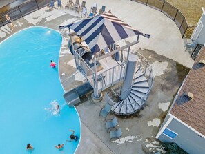 Une piscine avec un toboggan et un escalier en colimaçon menant à un espace ombragé équipé de chaises.