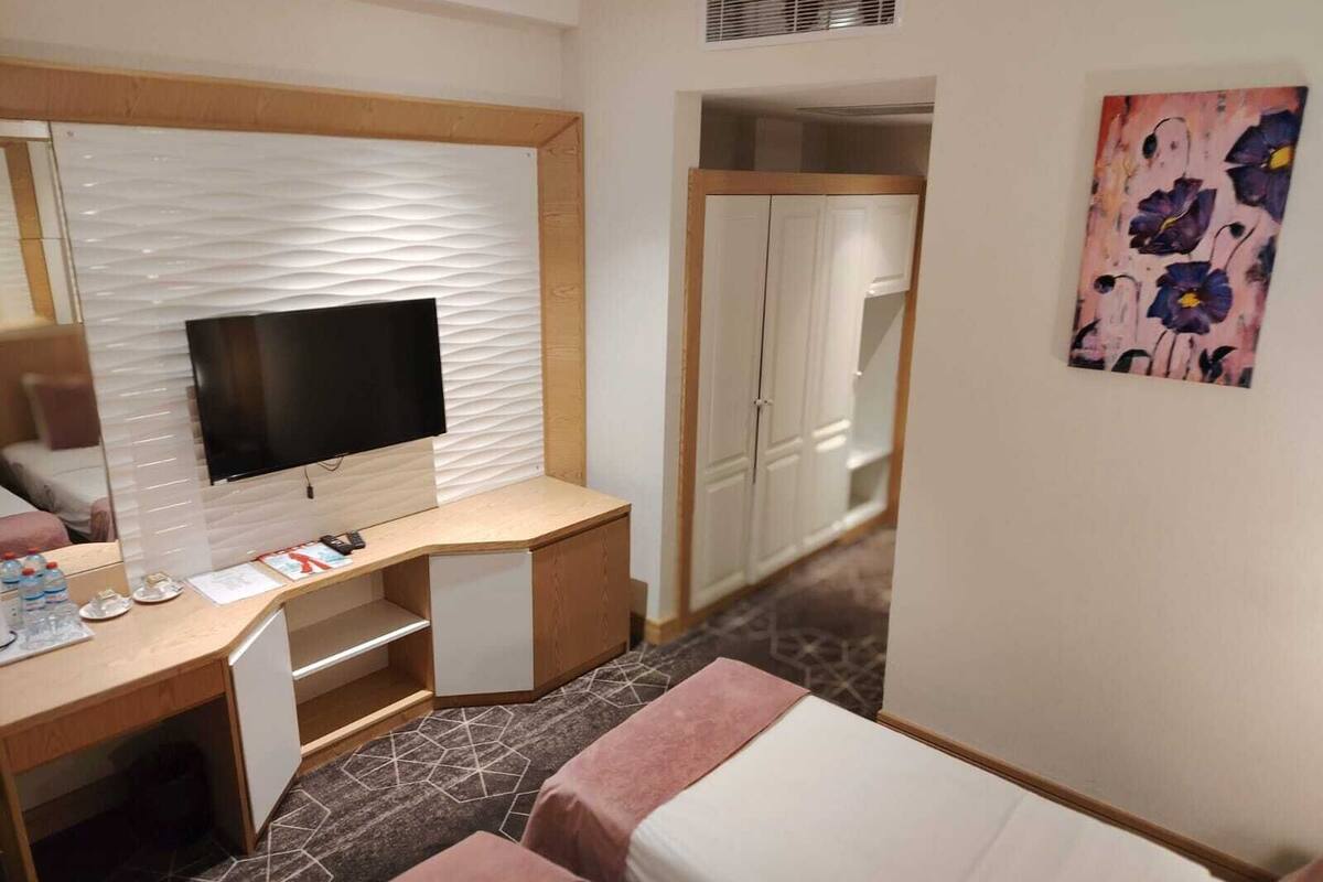 A hotel room with a bed, a desk with a flat-screen TV, a wardrobe, and a painting on the wall.