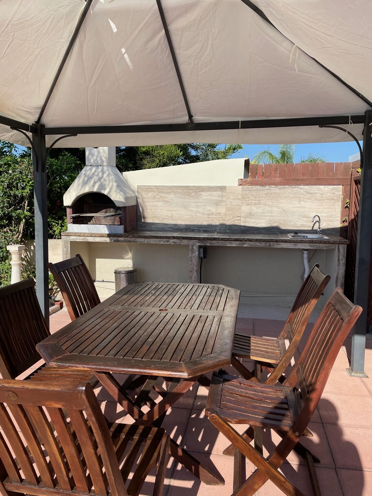 An outdoor patio with a wooden table and chairs, a pizza oven, and a sink under a canopy.