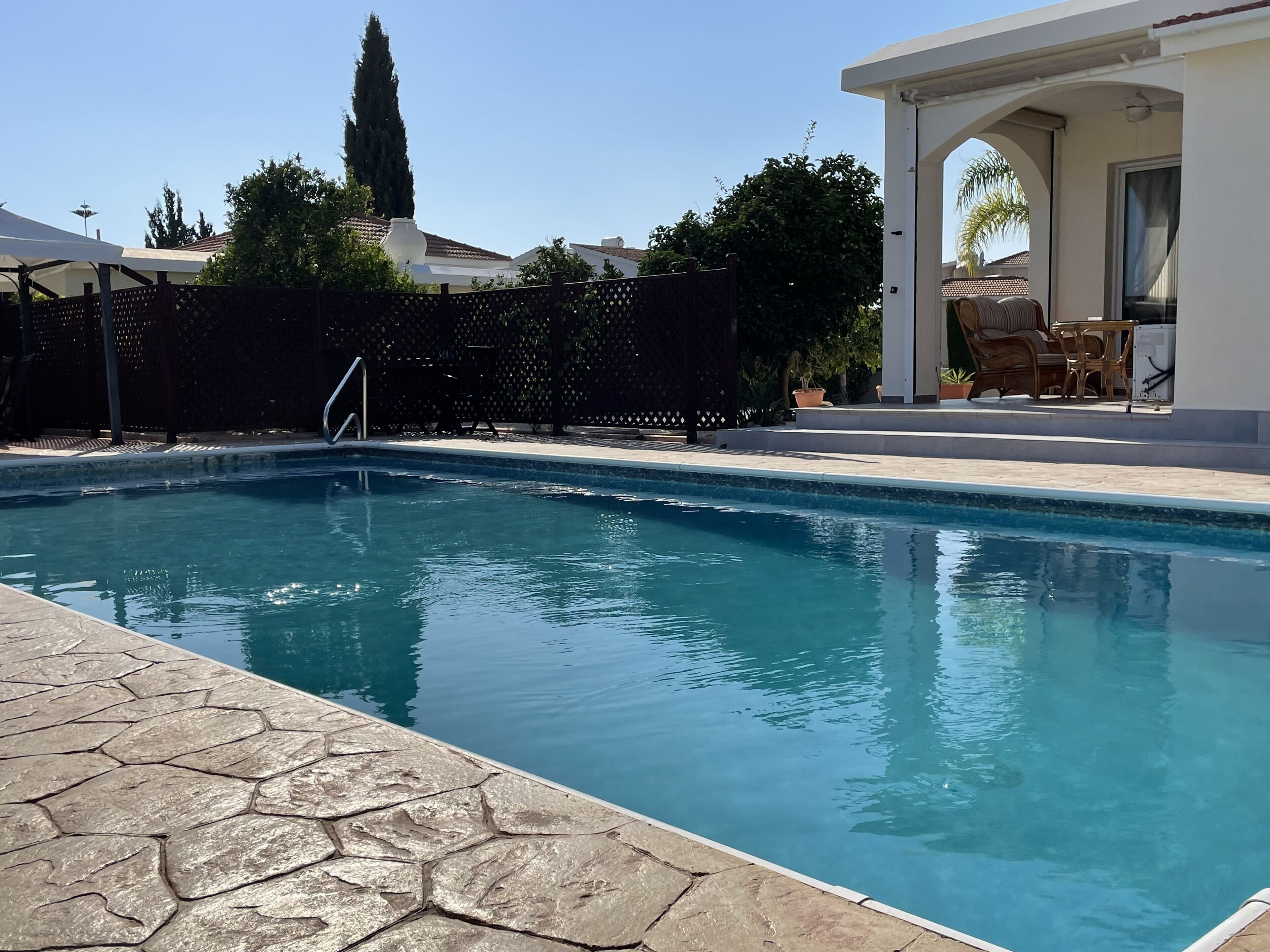 A swimming pool with stone tiles, a covered patio area, and a view of a tall tree and buildings in the background.