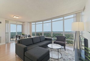 Ein modernes Wohnzimmer mit einem grauen Sofa, einem runden Glastisch und Blick auf eine Skyline durch große Fenster.