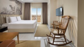 A hotel room with a large bed, a wooden rocking chair, a desk, a TV, and a balcony with a view. - Vibotel Residence Hotel Apartments (New Cairo)