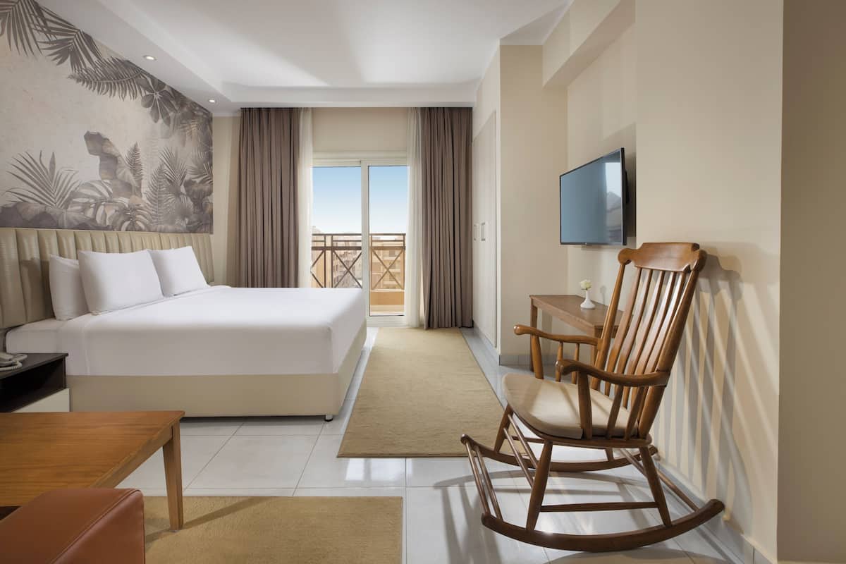 A hotel room with a large bed, a wooden rocking chair, a desk, a TV, and a balcony with a view.